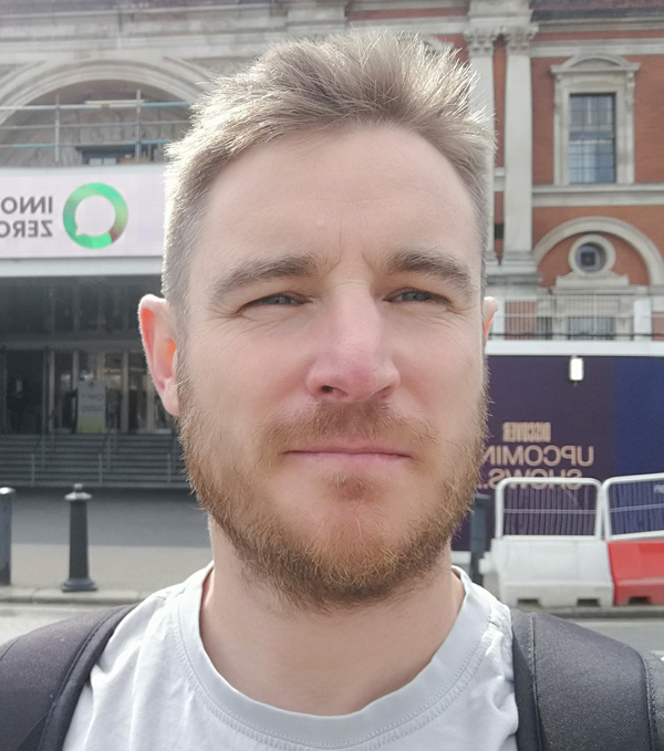 A young man with blond hair and beard taking a selfie outdoors city background.